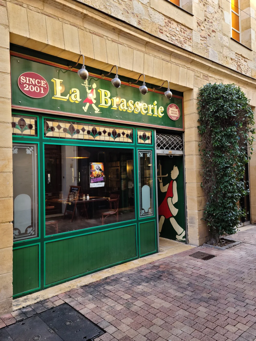La Brasserie restaurant in Bergerac with a bright green facade, stained-glass windows, and ivy-covered stone wall beside cobblestone paving