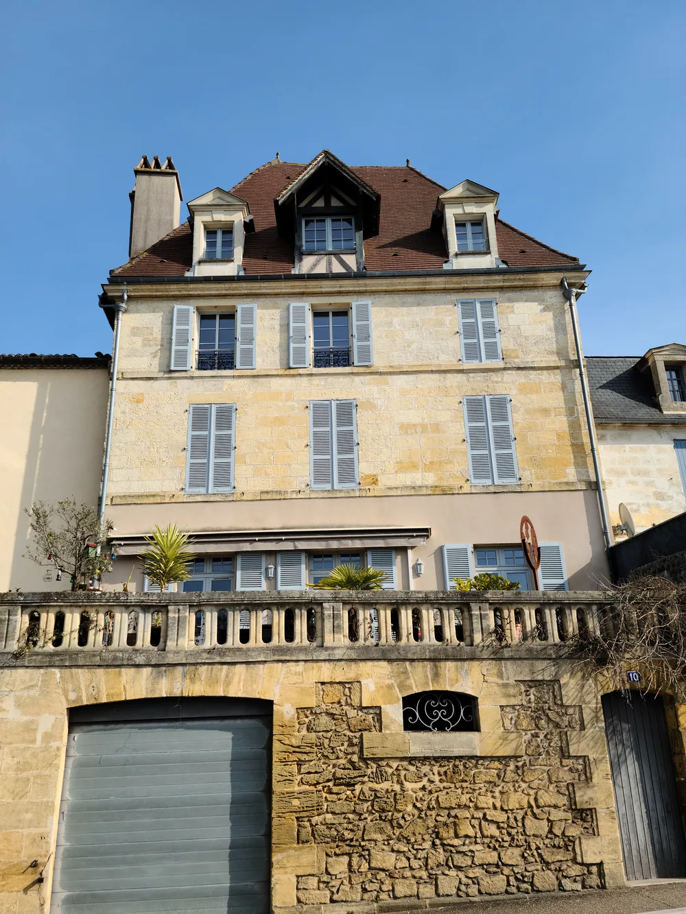 Multi-storey stone building in Bergerac with layered shuttered windows, a rooftop terrace with plants, and a contemporary restaurant at street level