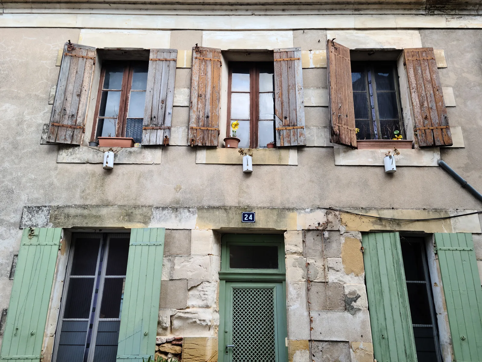 Weathered stone building facade in Bergerac with aged wooden shutters, faded plaster, and small sunflower pots on window ledges