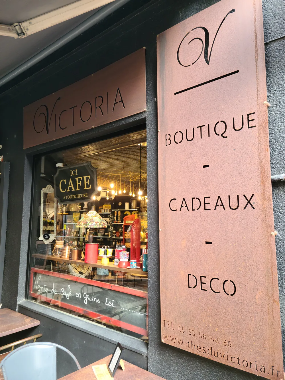 Victoria boutique sign in rose-toned metal on a shopfront in Bergerac, advertising boutique, gifts, and decor beside an outdoor cafe table