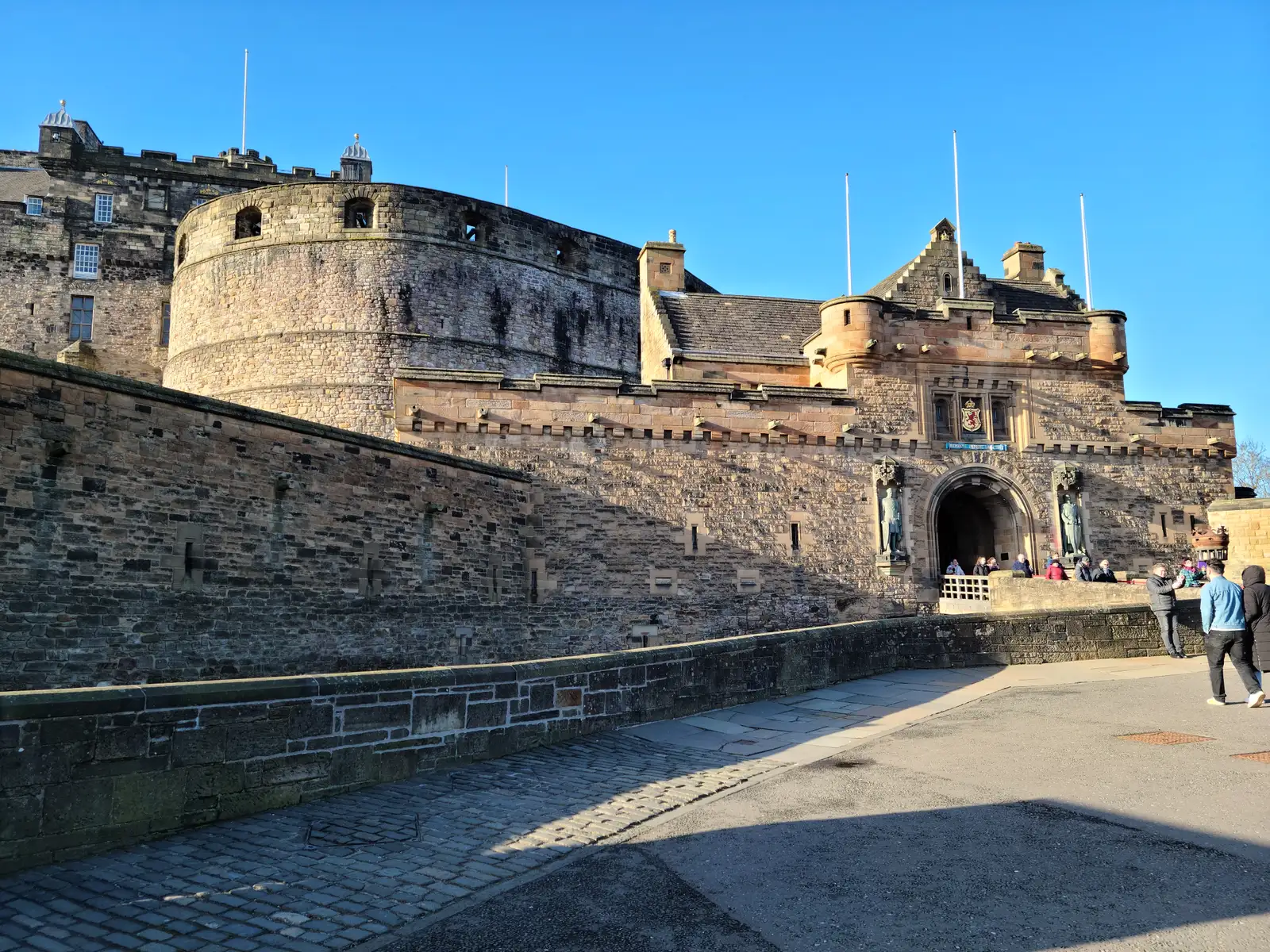 Edinburgh Castle's gatehouse with arched portcullis entrance, statues of Bruce and Wallace, and visitors crossing the esplanade bridge