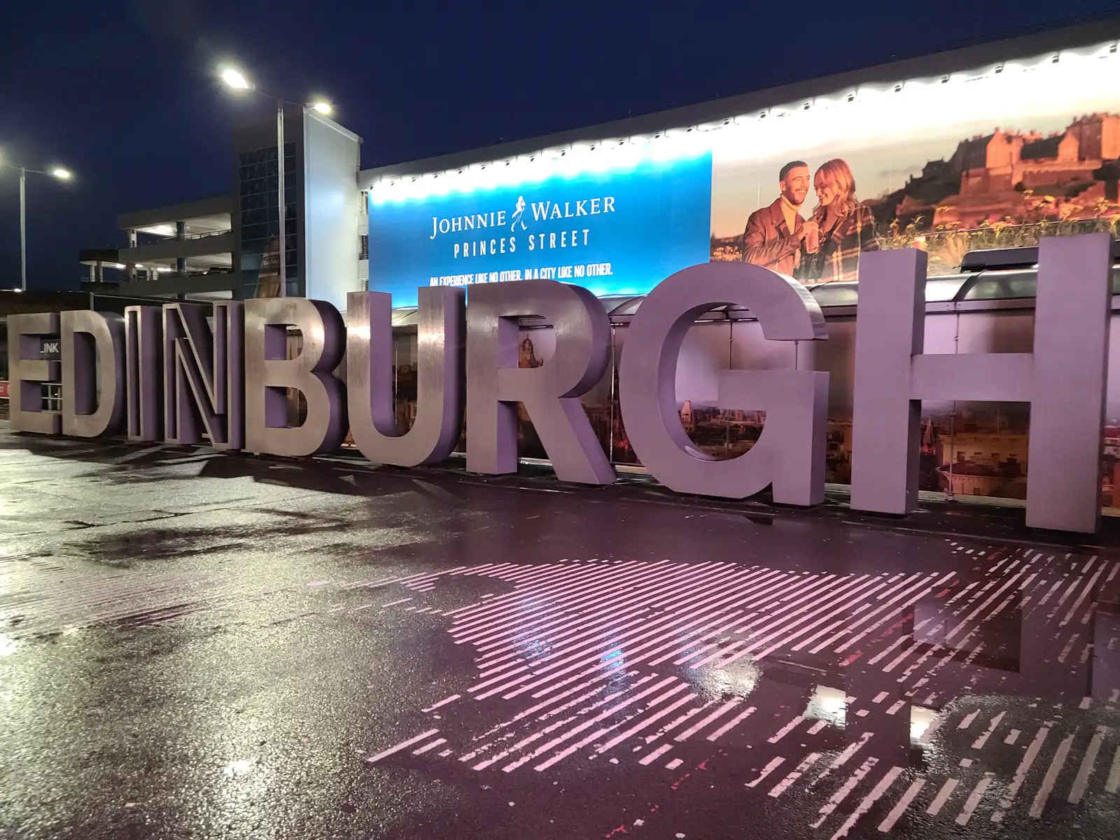 Illuminated giant Edinburgh letter sculpture outside the airport at night with Johnnie Walker Princes Street billboard behind