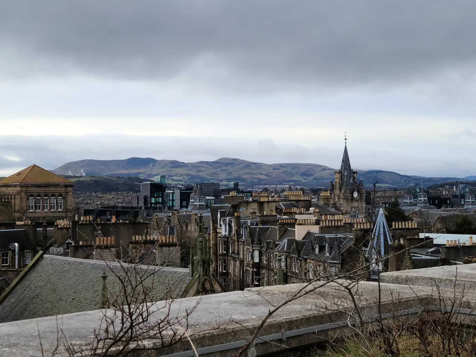 View from Calton Hill over Edinburgh Old Town rooftops with a clock tower spire and the Pentland Hills under cloudy skies