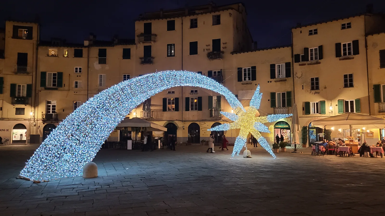 Piazza dell'Anfiteatro in Lucca at night with a sweeping blue and gold Christmas light installation arching over the oval square, restaurants lit in the background